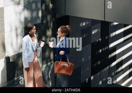 Smiling businesswomen standing on stairs outdoors with take out coffee and gossiping about work Stock Photo