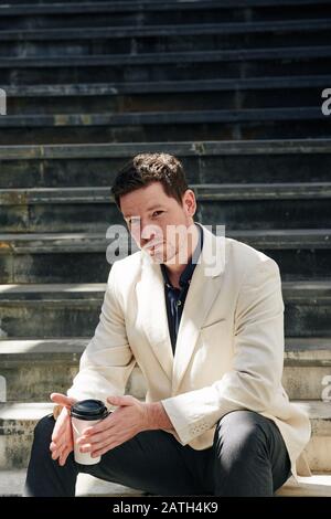 Portrait of young handsome man resting on steps and drinking take out coffee Stock Photo