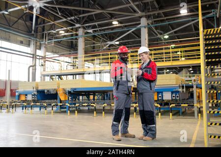 Young interracial colleagues in protective workwear using portable device while planning production volume at factory Stock Photo