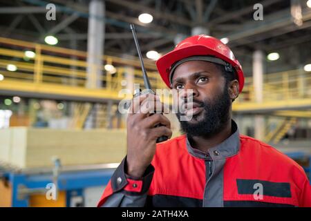 Young bearded Black laborer in red uniform transferring message to coworker with walkie-talkie device at construction site Stock Photo