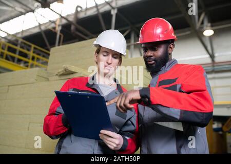 Below view of bearded Black man in hardhat asking colleague about industrial plan while pointing at paper in clipboard Stock Photo