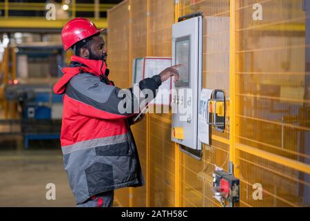 Serious young Black man in workwear and helmet choosing setups on control panel in factory shop Stock Photo