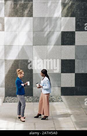 Young businesswomen standing outdoors, drinking take away coffee and discussing fresh news and ideas Stock Photo