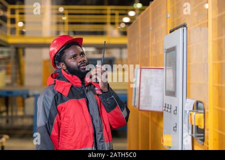 Serious bearded Black repairing engineer in hardhat using walkie-talkie while reporting on machine process Stock Photo