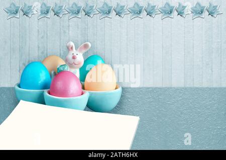 Easter eggs in a rabbit-shaped stand and a blank sheet of paper . Monochrome blue background with bells Stock Photo