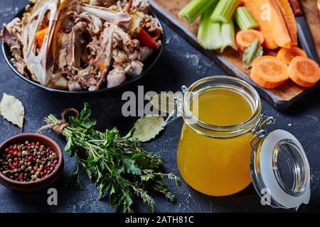 cooked chicken stock in a glass jar on a kitchen table with bouquet garni and chicken bones in a bowl, horizontal view from above Stock Photo