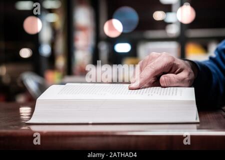 Close up view of male hand on the book. Man reading a book while sitting at a table in a library or book store. Selective focus on hand, blurred background. Stock Photo