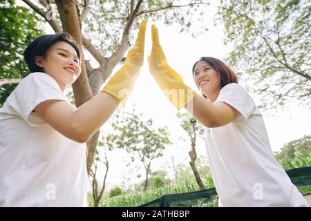 Happy young female volunteers in rubber gloves giving each other high five after finishing collecting garbage in city park Stock Photo