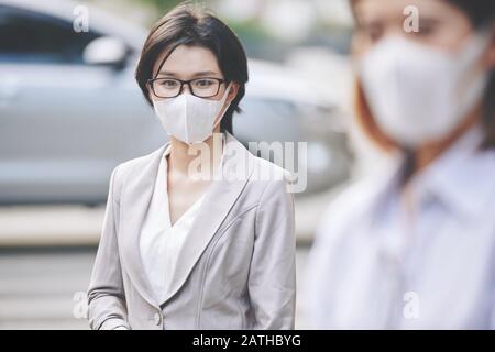 Portrait of young Asian business lady on glasses wearing face mask when walking in city to prevent getting dangerous virus Stock Photo