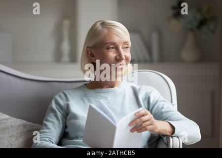 Happy mature woman distracted from reading book at home Stock Photo
