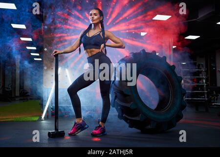 Side view of muscular woman holding big sledgehammer near tire wheel, looking away. Portrait of female bodybuilder in sportswear posing in gym, having rest after hard training. Concept of sport. Stock Photo
