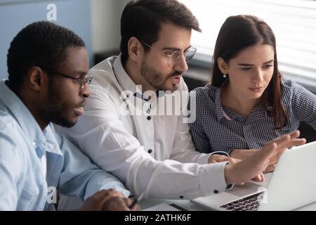 Three diverse colleagues looking at laptop screen working together indoors Stock Photo