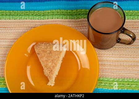 Slice of home-made coconut cake on orange coloured plat next to glass cup of milky tea standing on striped cloth table mat Stock Photo