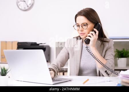 Serious attractive young secretary in glasses answering phone call while searching for information on laptop in office Stock Photo