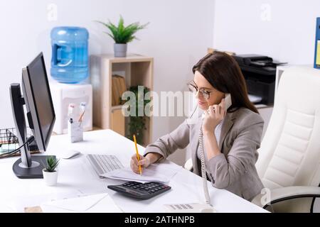 Friendly young female office manager in glasses checking documents while talking to customer by telephone Stock Photo