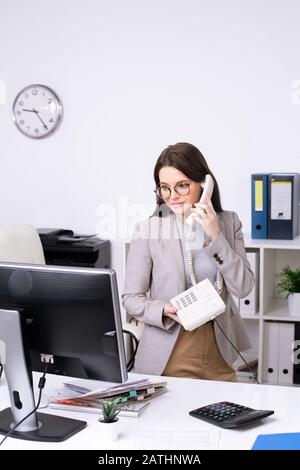 Content attractive young businesswoman in glasses standing at desk and talking on phone in office Stock Photo