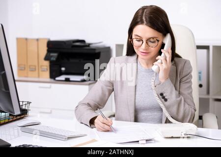 Confident young businesswoman sitting at table and making notes in document while discussing contract content by phone Stock Photo