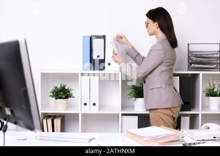 Brunette secretary in jacket cleaning up office room and sorting files in folders Stock Photo
