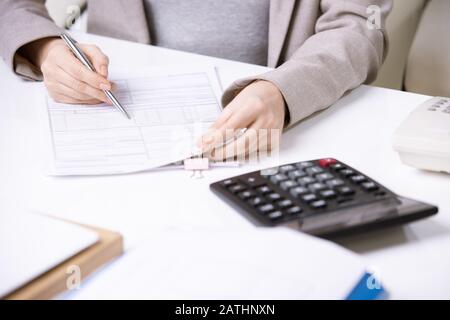 Close-up of unrecognizable office woman in jacket sitting at table with calculator and filling financial form Stock Photo