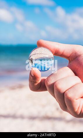 Person's Hand Holding Mini Glass Ball and Beach Background Stock Photo