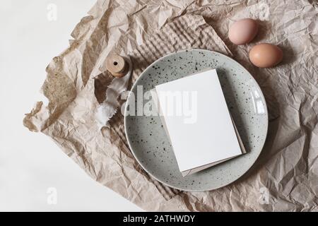 Spring breakfast scene. Lifestyle mockup composition with blank greeting card, ceramic plate and chicken eggs on brown craft paper. White table backgr Stock Photo