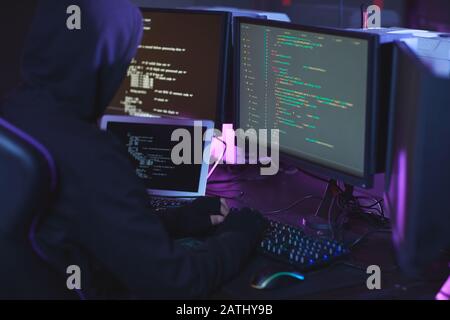 Back view of unrecognizable cyber security hacker wearing hood while working on programming code in dark room, copy space Stock Photo