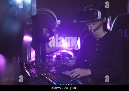 Side view portrait of young Asian man wearing VR headset while playing video games using racing shift in dark cyber interior, copy space Stock Photo