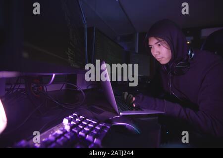 Asian cyber security hacker wearing hood while working on programming in dark room, copy space Stock Photo