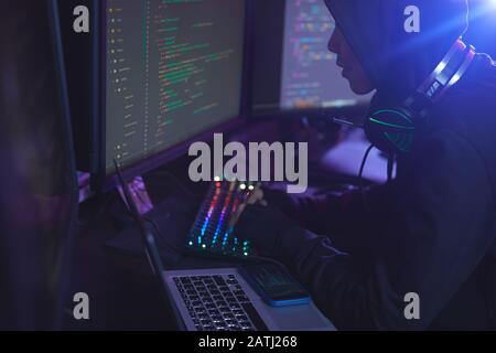 Side view at unrecognizable cyber security hacker wearing hood while working on programming in dark room, copy space Stock Photo