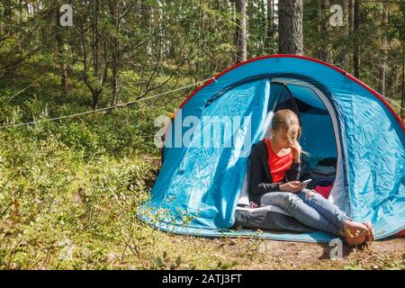 Tween girl sitting alone in camping tent focused on her smartphone messaging or checking social media during summer holidays Stock Photo