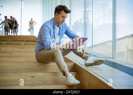 Man using tablet, sitting on steps at big windows, big working space. Stock Photo