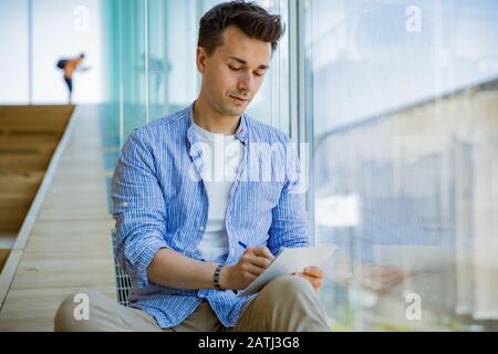 Man writing in notebook, sitting on steps at big windows, big working space. Stock Photo
