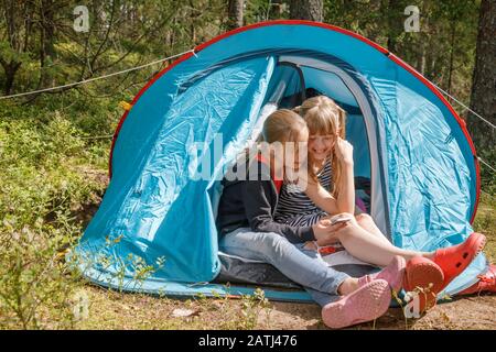 Tween girls sitting together in a camping tent using smartphone messaging or checking social media in a summer forest during summer holidays Stock Photo