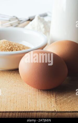 Baking preparation scene with two brown eggs in foreground, sugar, jug, flour and whisk in background on wooden kitchen chopping board. Filtered for r Stock Photo