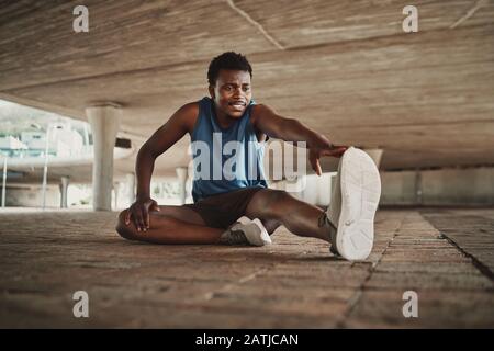Smiling portrait of a young man stretching his arms and legs while sitting on pavement under the bridge Stock Photo