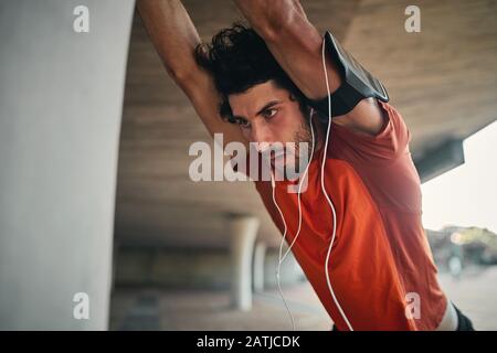 Exhausted sporty serious young man in earphone and armband warming up before running on street