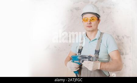 Portrait man builder repairer in protective glasses, hard hat, overalls showing having equipment in arm on grey background home brick banner Stock Photo
