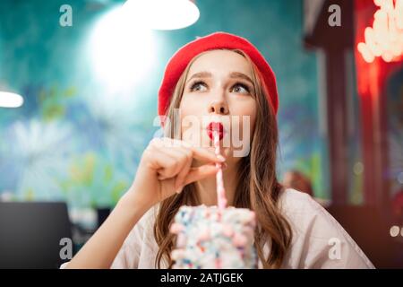 Girl with white teeth, beautiful smile in red beret, France drinks pink strawberry milkshake from straw Stock Photo