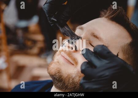 Acne removal with cosmetic product, cleaning pores of men skin, barber applying black charcoal mask to client man in spa salon Stock Photo