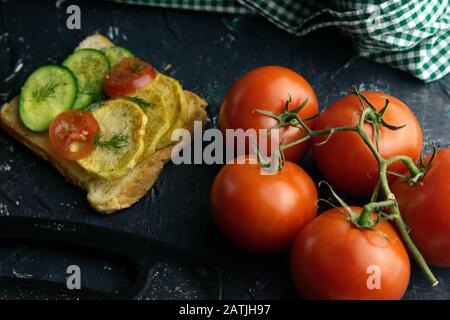 Sandwich with potatoes, cucumber and tomato next to tomatoes on a branch, stand on a black table. Stock Photo