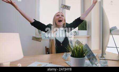 Elegant blond female sitting at desk at workplace with computer holding in hands bundle of cash looking astonished at monitor, and throwing money in air and happily making yes gesture. Stock Photo