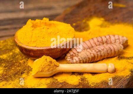 Dried curcuma powder in a wooden bowl with a wooden spoon and whole fresh turmeric roots in front of a wooden background. Stock Photo