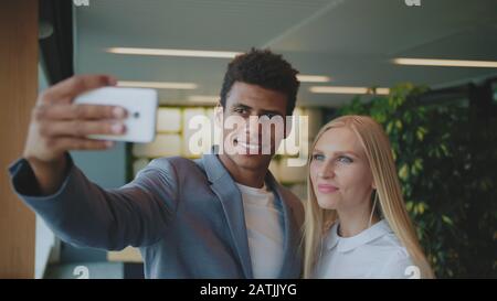 Cheerful black man with laughing blond woman taking selfie with smartphone in modern office having fun. Stock Photo