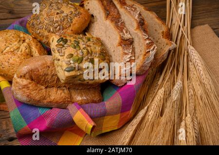 Freshly various buns and bread slices in a basket and a half whole grain bread on a jute fabric placed on a rustic wooden background with grain ears. Stock Photo