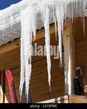 Wooden house with snow cornice and big icicles on roof and ski equipment on balcony at sun winter day Stock Photo