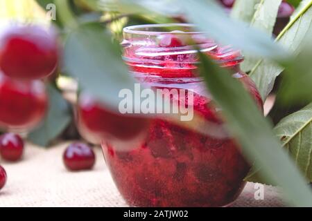 Cherry jam in a glass jar on a background of green leaves on a table of wooden boards. Close-up. Stock Photo