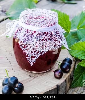 Black currant jam in a glass jar on a wooden table near green leaves and currant berries. Natural currant jam. Close-up. Stock Photo