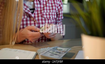 Crop view of blond female in checkered casual shirt, sitting at wooden table with dollar cash on it turning bitcoin over in hands studying it Stock Photo