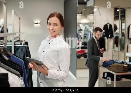 Portrait of smiling beautiful young saleswoman in shirt standing at rack and checking receiving merchandise in clothes shop Stock Photo