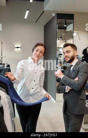 Friendly clothing store consultant in blouse demonstrating new collection of clothes while working with client in shop Stock Photo